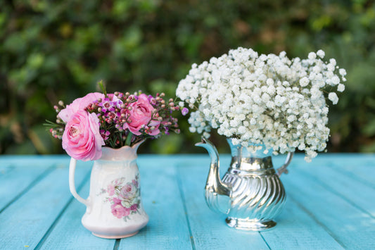Vintage tablescape décor featuring pink ranunculus in a floral ceramic pitcher and white baby’s breath in a silver teapot vase on a turquoise table—perfect for cottagecore and shabby chic styling.