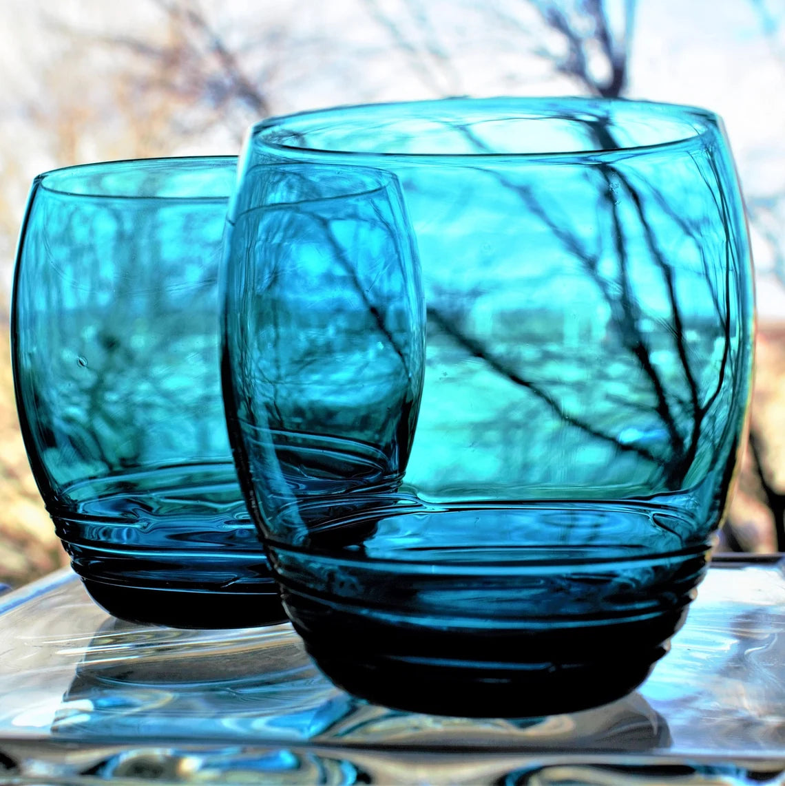 Two blue glass tumblers on a reflective surface with blurred natural background