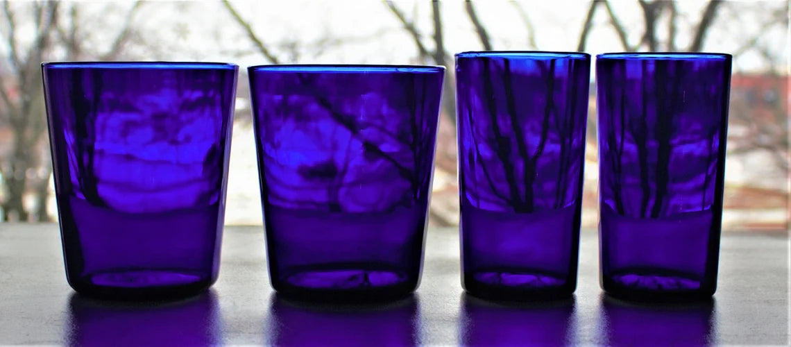 Four cobalt blue glass tumblers on a reflective surface with a blurred outdoor background