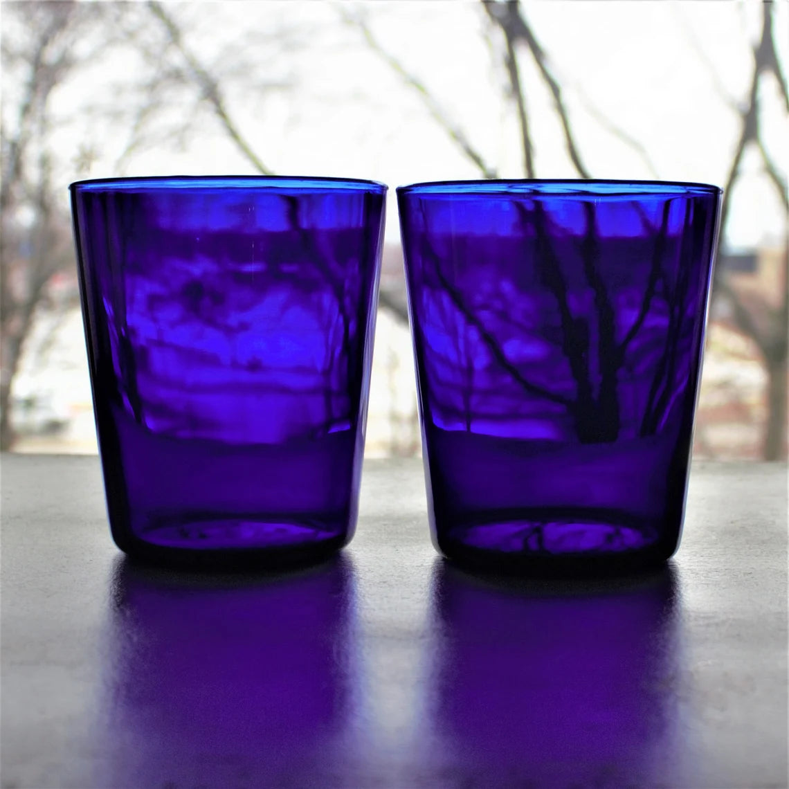 Two cobalt blue glass tumblers on a reflective surface with a blurred natural background