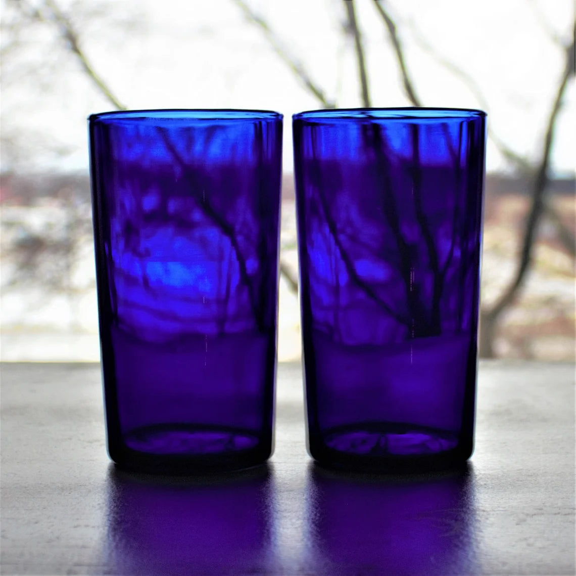 Two cobalt blue glass tumblers on a reflective surface with a blurred outdoor background