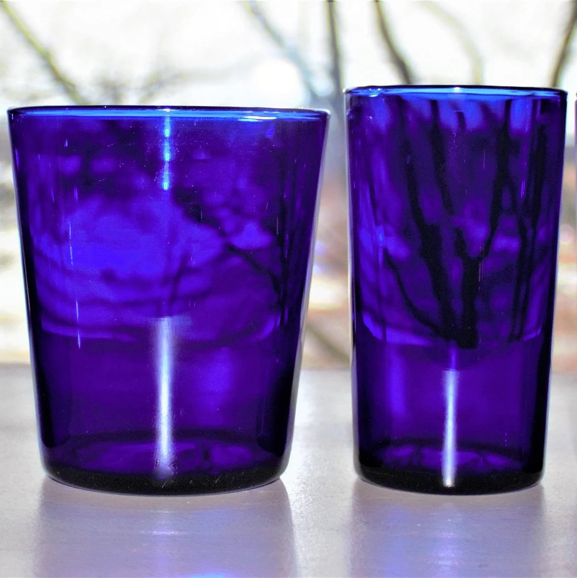 Two cobalt blue glass tumblers on a reflective surface with a blurred natural background