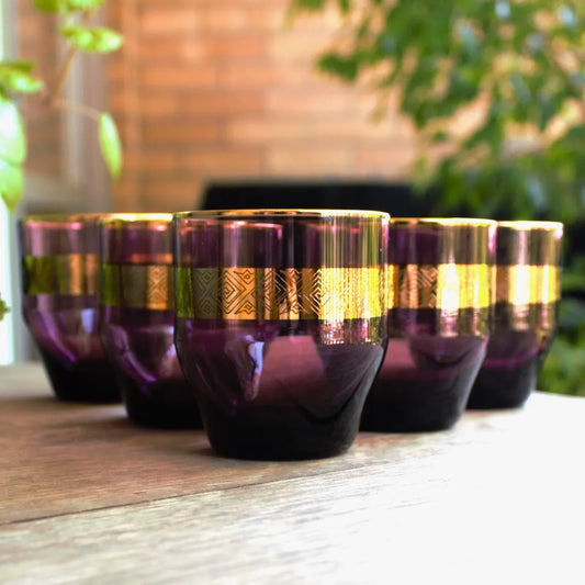 Set of purple glass tumblers with gold accents on a wooden surface outdoors.