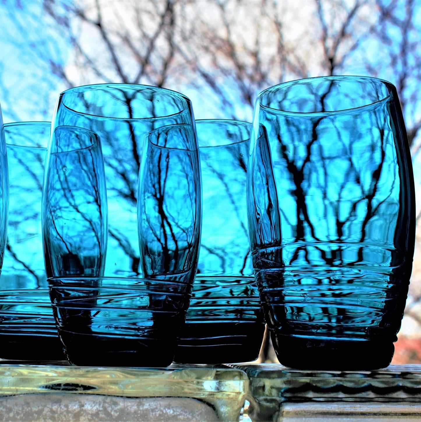 Set of blue glass tumblers on a reflective surface with blurred natural background