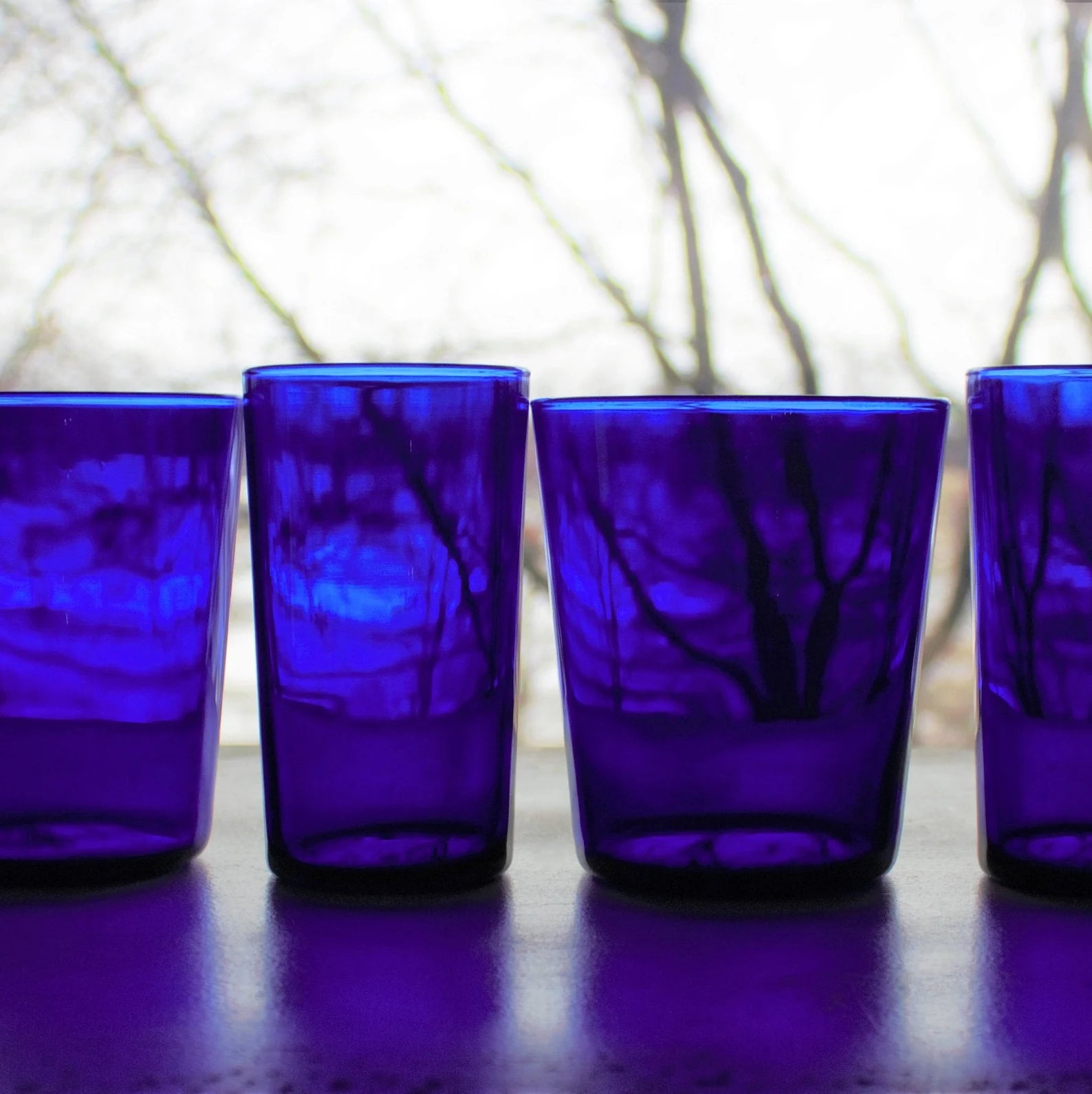 Four cobalt blue glass tumblers on a reflective surface with a blurred natural background