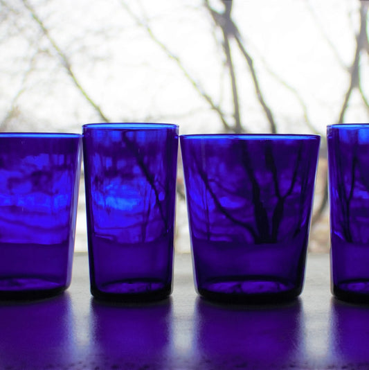 Four cobalt blue glass tumblers on a reflective surface with a blurred natural background